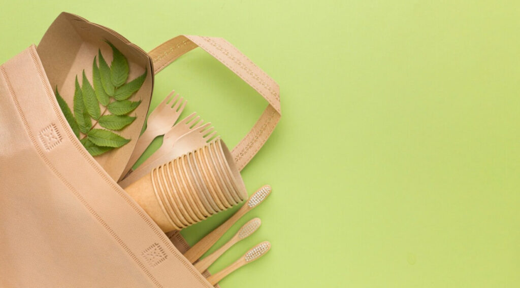 beige eco bag with forks, toothbrushes, cups, and green leaf on green background